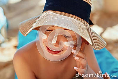 Facial Care. Female Applying Sun Cream and Smiling. Beauty Face. Portrait Of Young Woman in hat Smear Moisturizing Lotion on Skin. Stock Photo