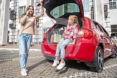 Fair-haired girl sitting in car boot, her mom talking on the phone Stock Photo