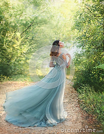 Fairy tale princess in light summer blue, turquoise dress standing in park. Happy Woman walks towards wind, graceful Stock Photo