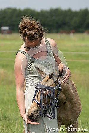 A falk color foal in the field, wearing a fly mask, young woman takes off the fly mask Stock Photo