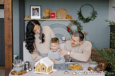 The family bakes a gingerbread house for Christmas with sweets, the father puts icing on his son`s nose, they laugh Stock Photo