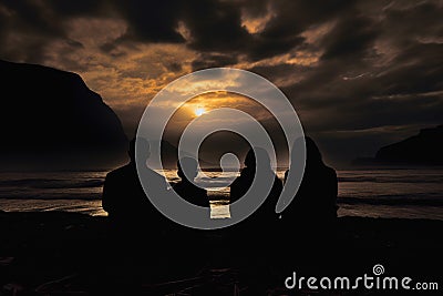 Family of four sitting on ocean shore and looking at dramatic orange sunset at dusk, rear view Stock Photo