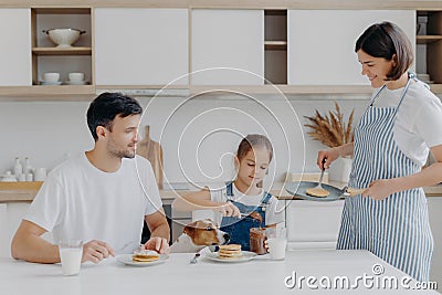 Family have breakfast at kitchen in morning. Happy girl puts melted chocolate on tasty fried pancake, poses at table with father, Stock Photo