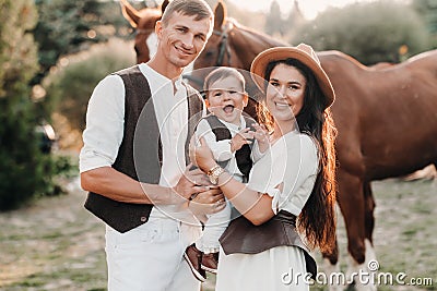 A family in white clothes with their son stand near two beautiful horses in nature. A stylish couple with a child are photographed Stock Photo