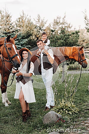 A family in white clothes with their son stand near two beautiful horses in nature. A stylish couple with a child are photographed Stock Photo
