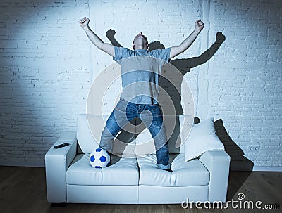 Fanatic and crazy football fan watching television soccer match with ball jumping on sofa celebrating goal Stock Photo