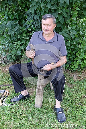 Farmer with hammer and iron tool on the tree stump is sharpening his scythe Editorial Stock Photo