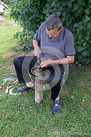 Farmer with hammer and iron tool on the tree stump is sharpening his scythe Editorial Stock Photo