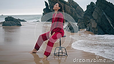 Fashion girl posing beach in red elegant suit overcast day. Woman sitting chair. Stock Photo