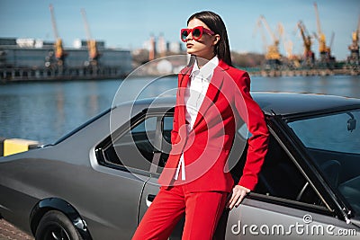 Fashion girl standing next to a retro sport car on the sun. Stylish woman in a red suit and sunglasses waiting near classic car Stock Photo