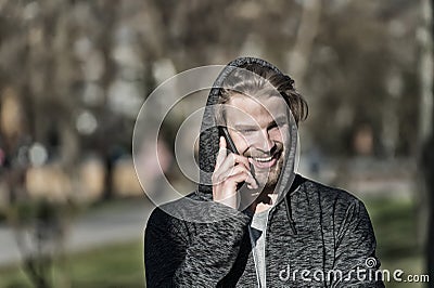 Fashion macho smiling with smartphone in casual sweatshirt. Happy guy in hood talk on mobile phone on sunny outdoor. Bearded man s Stock Photo