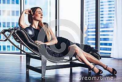 Fashion model with slim long legs wearing black cocktail dress lying on lounge chair in penthouse apartment Stock Photo