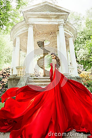 Fashion Model Waving Long Fluttering Red Dress, Woman in Garden, Old White Alcove in Flowers Stock Photo