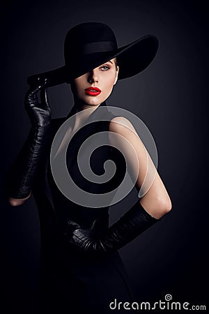 Fashion Model in Wide Brim Hat with Red Lips Make up. Elegant Retro Woman in Black Suit Dress and Gloves over Dark Gray Studio Stock Photo