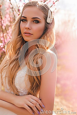 Beautiful girl with blond hair in elegant wedding dress posing among flowering peach trees in garden Stock Photo