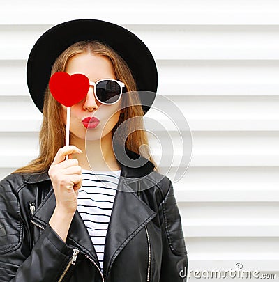 Fashion portrait face pretty sweet young woman with red lips making air kiss with lollipop heart wearing black hat leather jacket Stock Photo