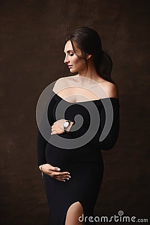 Fashion portrait of a gorgeous pregnant model girl wearing black evening dress posing over brown background Stock Photo