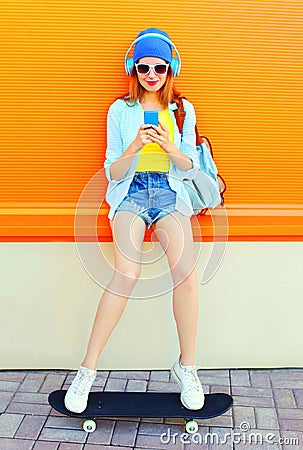 Fashion pretty cool girl is listens to music and using a smartphone sits on a skateboard over colorful orange Stock Photo