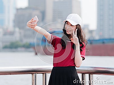 Fashion pretty young girl with black long hair, wearing red T-shirt and white baseball cap selfie with scissors hand pose outdoor Stock Photo