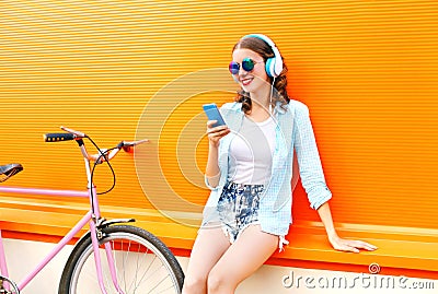 Fashion pretty young woman listens to music using smartphone near urban retro bicycle over colorful orange Stock Photo