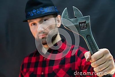 A fashionable car service employee in a red shirt and a black hat holds a large adjustable wrench in his hand on a black Stock Photo