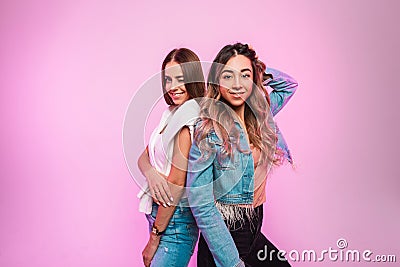 Fashionable joyful women in stylish casual jeans clothes stand with their backs to each other and smiling indoors. Pretty happy Stock Photo