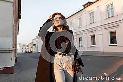 Fashionable joyful young hipster woman in a long coat in dark sunglasses in a t-shirt in white jeans with a leather bag Stock Photo