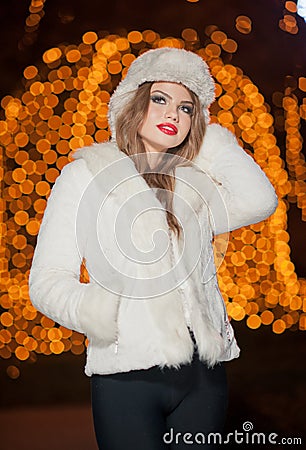 Fashionable lady wearing white fur cap and coat outdoor with bright Xmas lights in background. Portrait of young beautiful woman Stock Photo