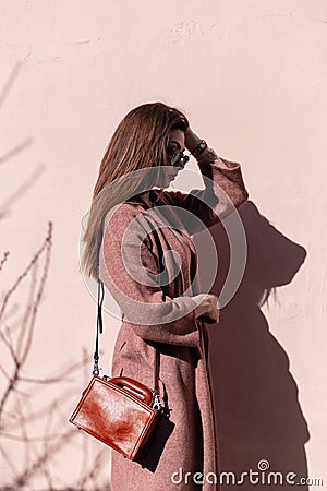 Fashionable pretty luxurious young woman model in stylish coat with handbag in sunglasses posing near vintage pink wall on sunny Stock Photo