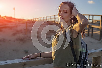 Fashionable slender young brunette girl with backpack posing on a background wooden walkways and sand beach at sunset Stock Photo