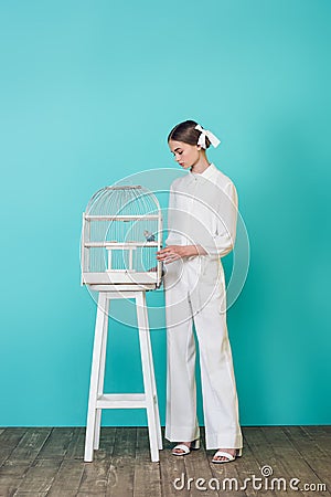 fashionable teen girl in stylish white outfit looking at parrot in cage Stock Photo