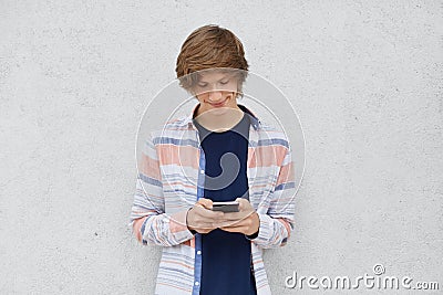 Fashionable teenage boy wearing shirt, holding cell phone in hands, messaging with friends or playing games online using free inte Stock Photo