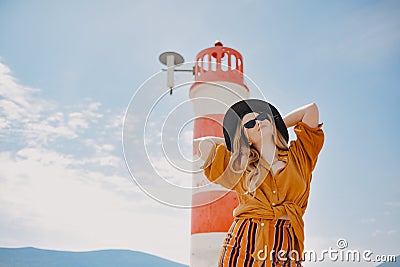 Fashionable woman on vacation against the backdrop of the sea, mountains and a lighthouse. Summer, sunny day, walk along the beach Stock Photo