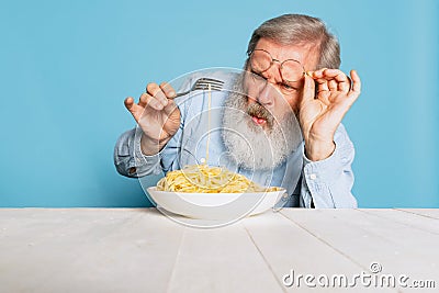 Fastidious senior hairy gray-bearded man tasting large portion of noodles, pasta on blue studio background. Stock Photo