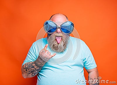 Fat man with beard, tattoos and sunglasses makes the gesture of the horns Stock Photo