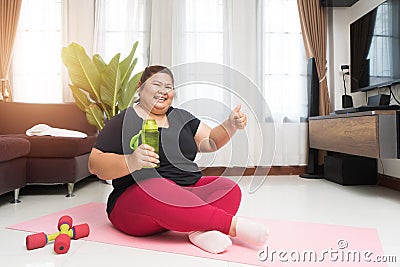 Fat woman asian holding a water bottle with showing thumbs up exercising at home, Stock Photo