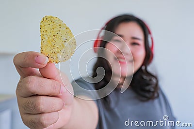 Fat woman holding potato chips indoors while listening music Stock Photo