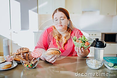 Fat young woman in kitchen sitting and eating food. Healthy lifestyle. Looking at burger and bowl salad. Body positive Stock Photo