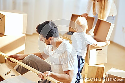 Father assembling furniture in living room of new apartment, mother with son and pile of moving boxes on background Stock Photo