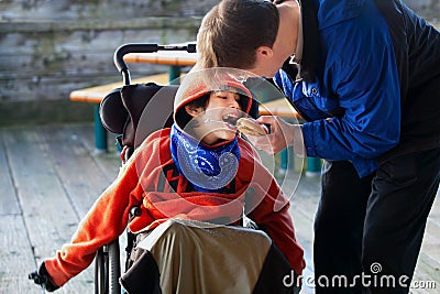 Father feeding disabled son a hamburger in wheelchair. Child has Stock Photo