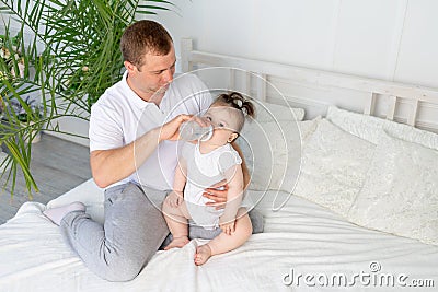 A father feeds his little daughter from a bottle on a white bed at home, the concept of baby food and child care Stock Photo