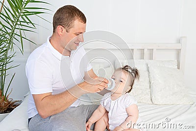 A father feeds his little daughter from a bottle on a white bed at home, the concept of baby food and child care Stock Photo