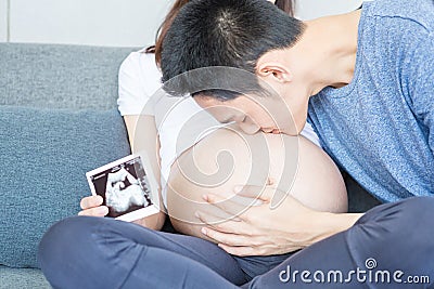Father is kissing his mother`s stomach. Young husband kissing his pregnant wife`s tummy in living room with the baby ultrasound Stock Photo