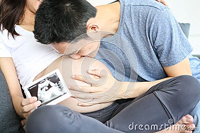 Father is kissing his mother`s stomach. Young husband kissing his pregnant wife`s tummy in living room with the baby ultrasound Stock Photo