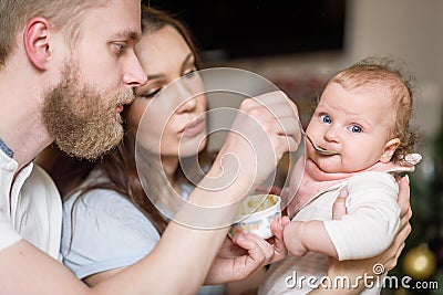 Father and mother feed their baby fruit puree in the kitchen from a spoon Stock Photo