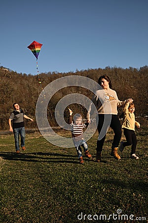 Father and mother walk with daughter and son on green grass at sunset and launch multicolored kite into sky. Have fun spending Stock Photo