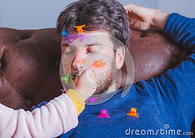 Father`s day. Young man sleeping while his little daughter paints his face Stock Photo