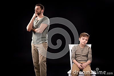 Father standing with hand on chin and upset little boy sitting on stool Stock Photo