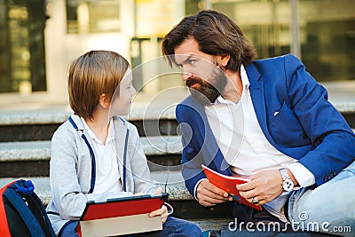 Father talking with son after lessons outside school. Schoolboy with backpack and notebooks. Dad and kid sitting at stairs before Stock Photo