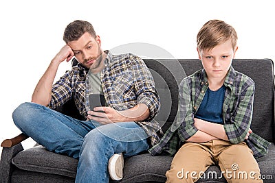 Father using smartphone and upset little son sitting on sofa with crossed arms Stock Photo